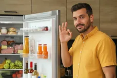 Hombre sonriente junto a un refrigerador abierto mostrando gesto de ok en la cocina